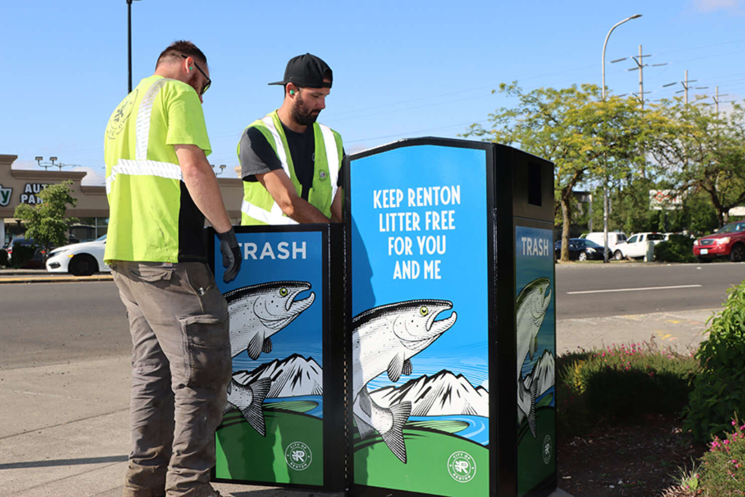 Two men in neon yellow coats emptying a trash receptacle with images of fish printed on it saying 