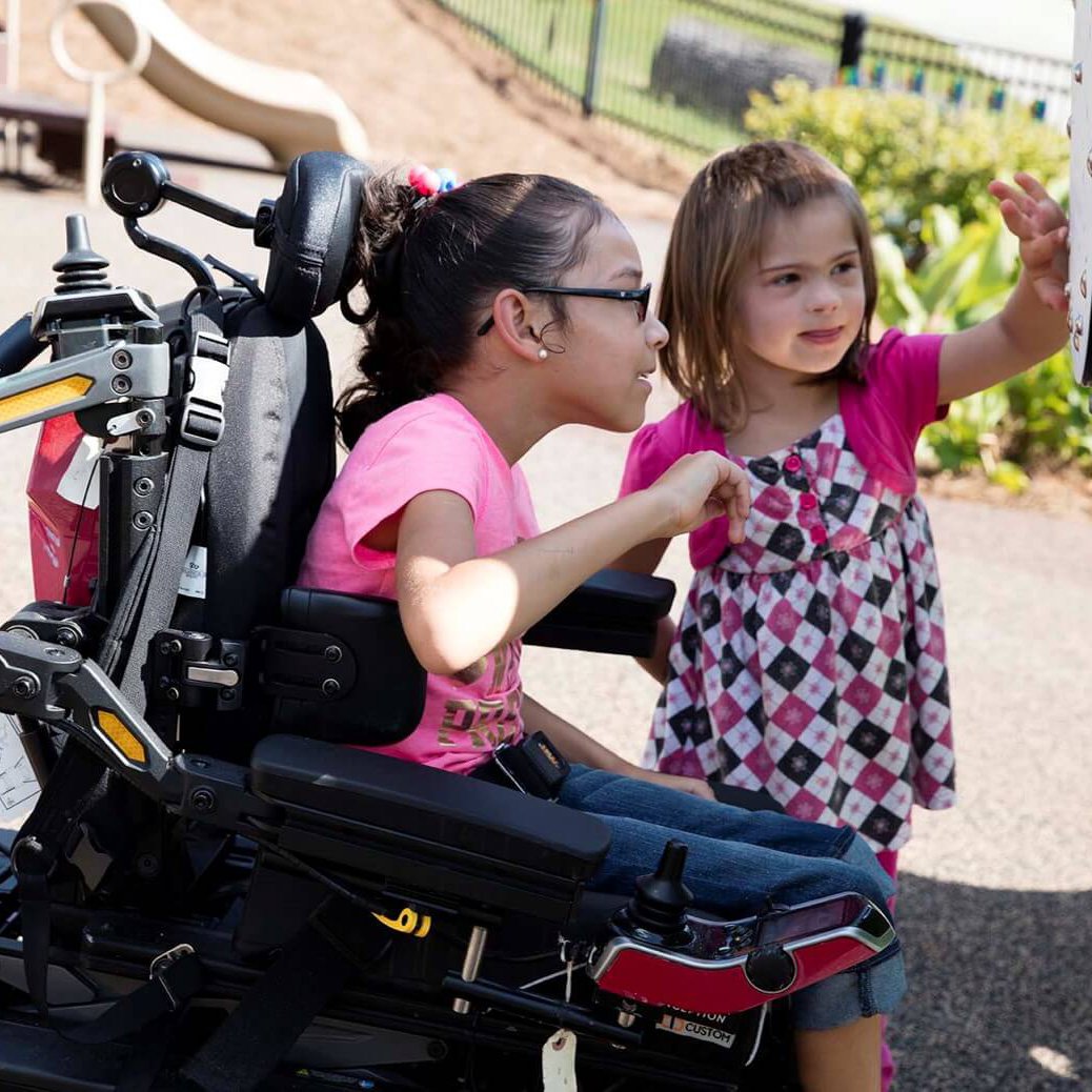 Two young girls, one using a wheelchair, engaging with an interactive play panel on an inclusive playground, promoting communication and accessibility.