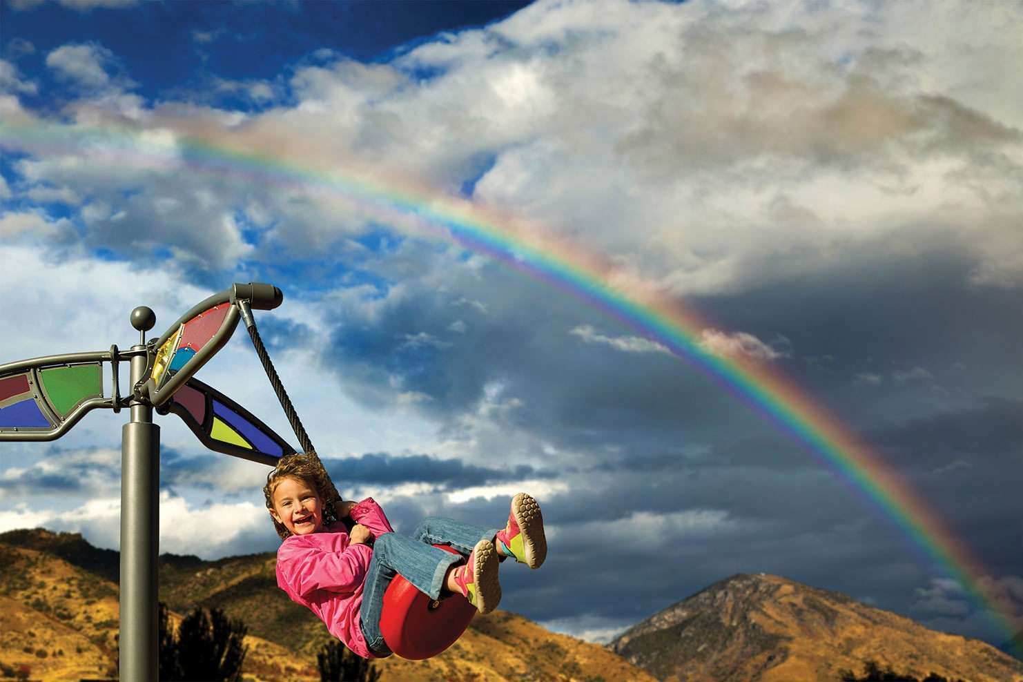 Image of a child in a pink coat playing on a swing with a rainbow in the background