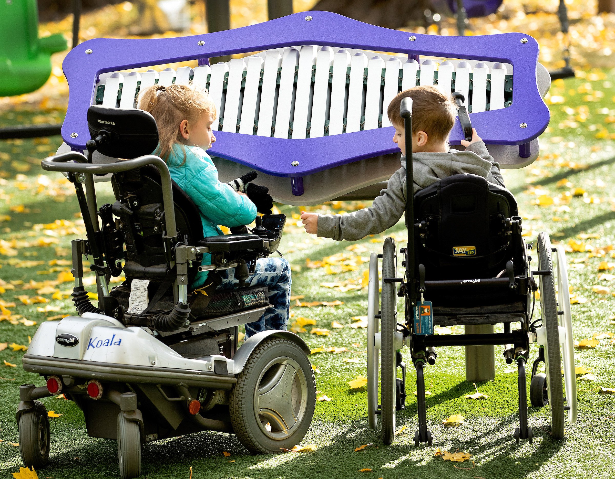 Two children in mobility devices playing with an outdoor musical instrument