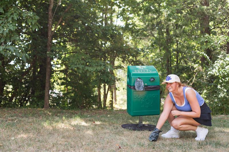 White woman with a blue shirt and black skirt is picking up waste with a black glove in front of a dog receptable