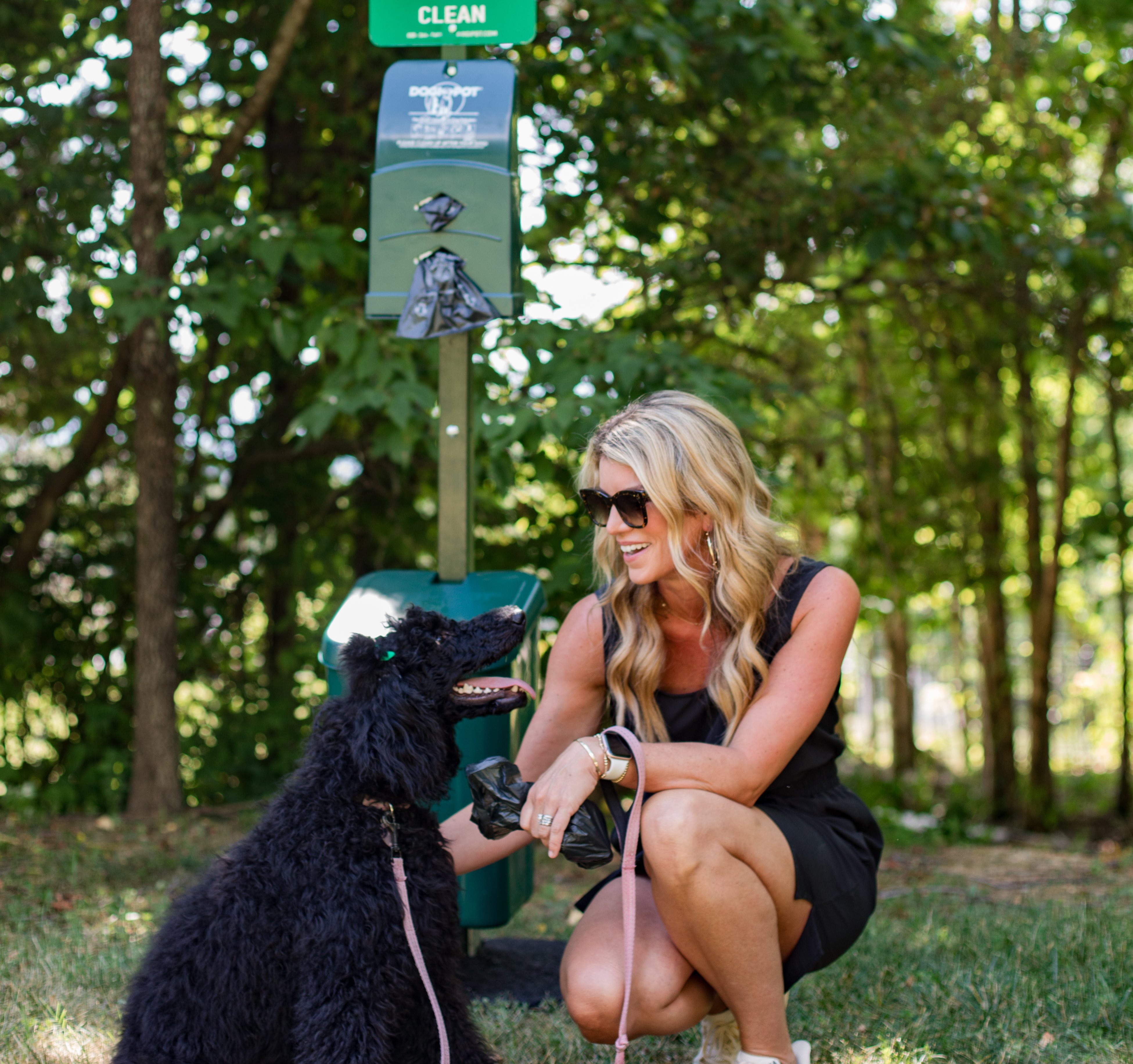Woman kneeling in front of DogiPot trash bin with black dog petting the dog.