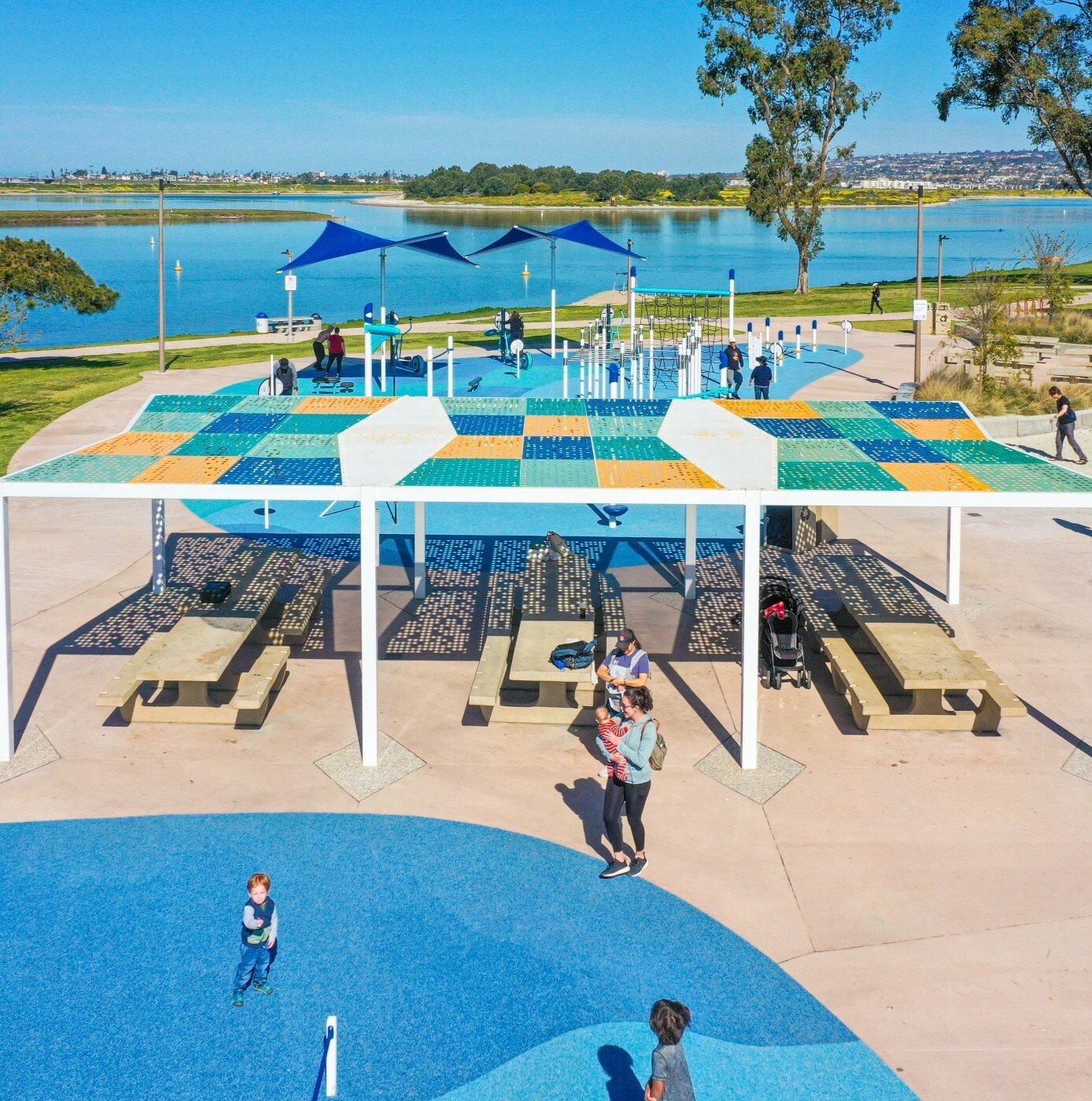 Image of people at a riverside playground that is predominately blue. In the image, there are picnic tables, shade features, and rubber designs present