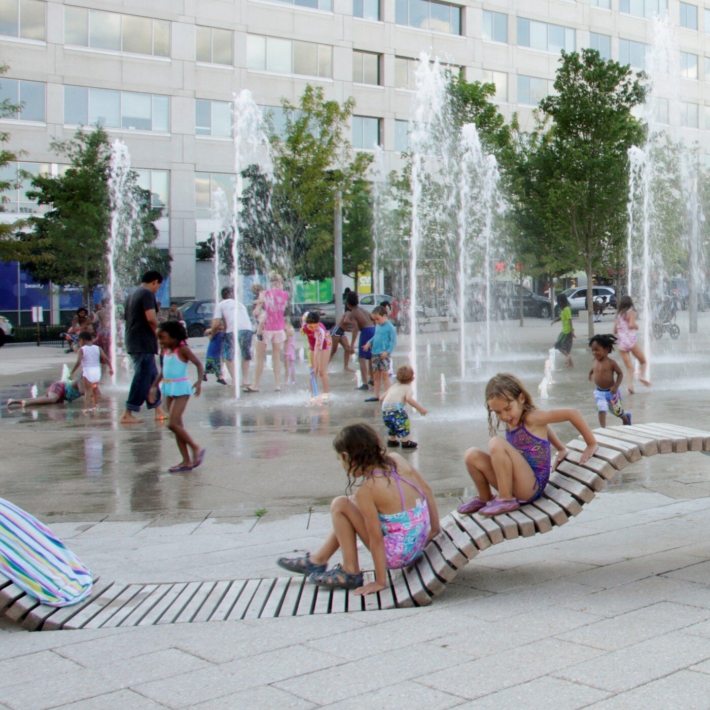 Image of groups of children playing in fountains at a water play park. There are also two children sitting on park seats as well