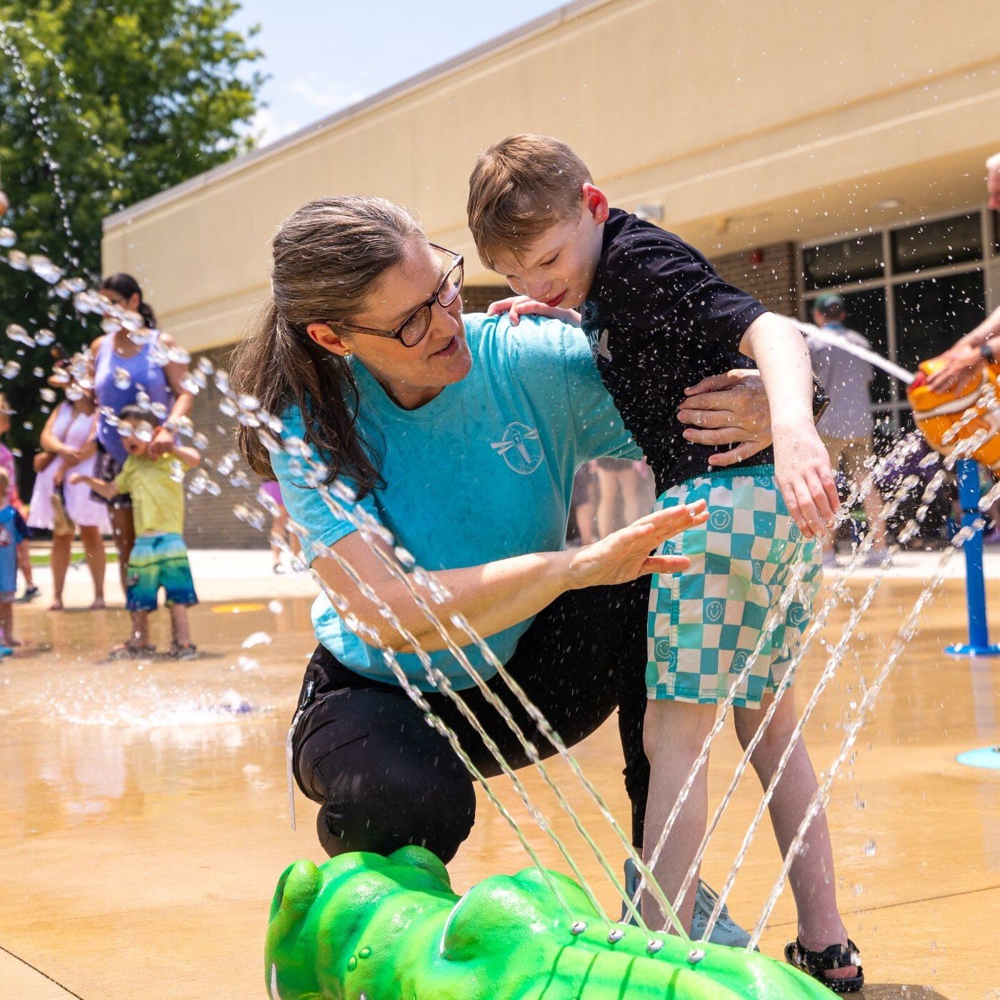 Image of a woman holding a child, while the child plays with the water coming from the green water fountain.