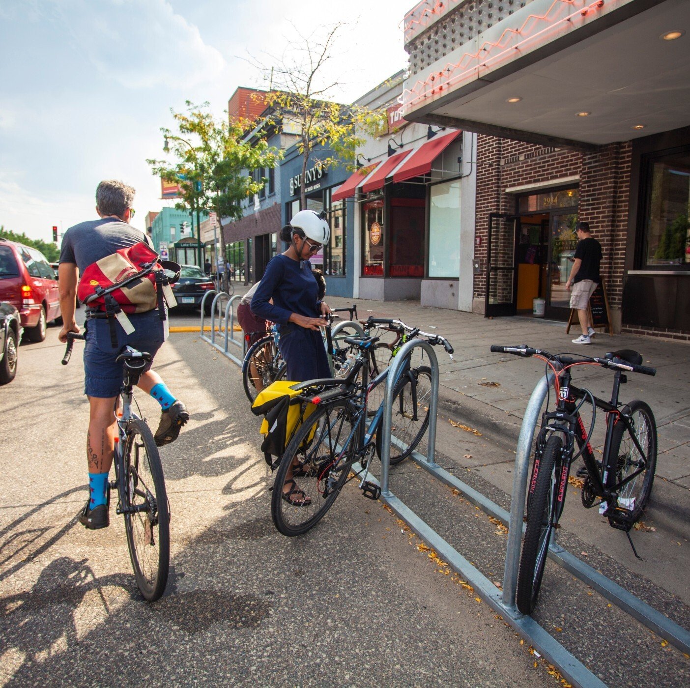 Image of a man on a bicycle riding away and a woman locking her bike onto a bike stand on the side of a street