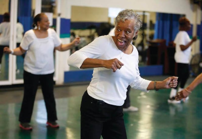 Older woman in a white top enthusiastically leads a dance-fitness move in an indoor exercise class, with other participants following in the background.