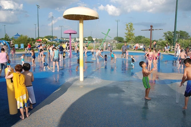 Children and adults play under mushroom-shaped sprayers and ground jets at a busy outdoor splash pad on a sunny day.