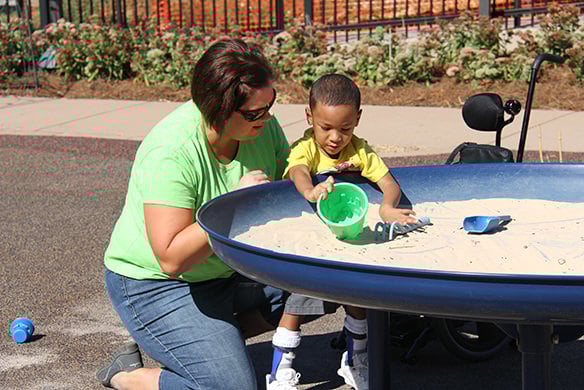 A lady and child play together in a circular table that contains sand. The child is seen scooping sand into a green sand bucket