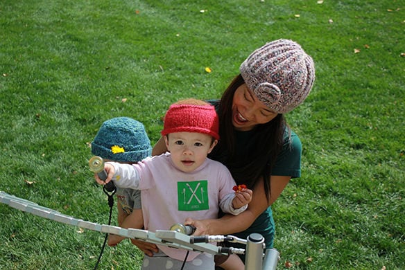 A baby in a pink shirt and red cap being held by a young lady in a grey beanie plays on outdoor musical equipment