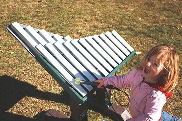 A young girl in a pink coat smiling as she plays with an outdoor xylophone