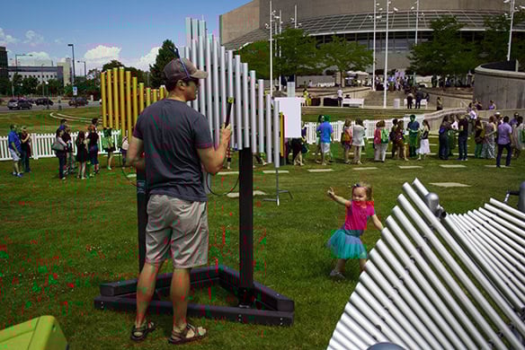 A young girl in a blue tutu and pink shirt is pointing at a middle-aged man in a grey shirt and shorts as he plays on the outdoor xylophone
