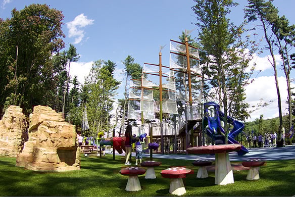 Playground that has a pirate ship, mushroom stools, and rocks, surrounded by trees and people.