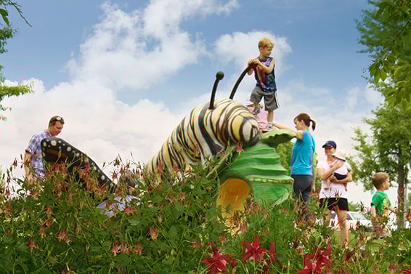 Playground with a large caterpillar sculpture that a child is standing on top of. The playground also contains people, flowers, and greenery nearby.