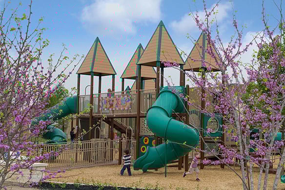 A playground with green and tan color accents that showcases two slides, a bridge, and trees