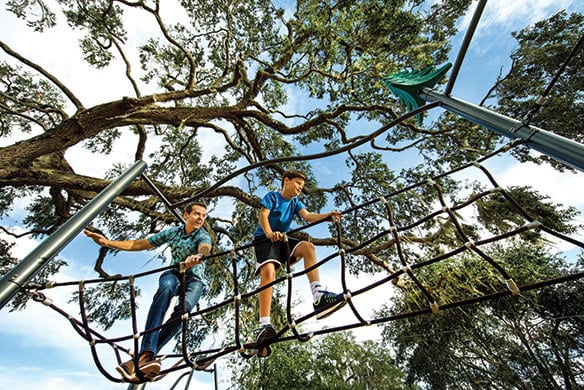 A man and teenager walk across a rope bridge with trees shown above them
