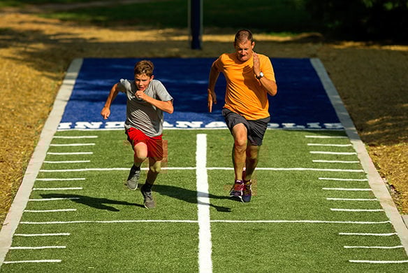 A young man in a grey shirt and an older man in a gold t-shirt race one another in a 40-yard dash