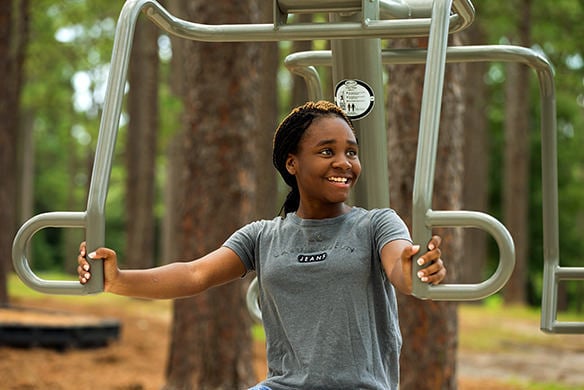 A teenage girl performing chest press on outdoor exercise equipment in a wooded area 