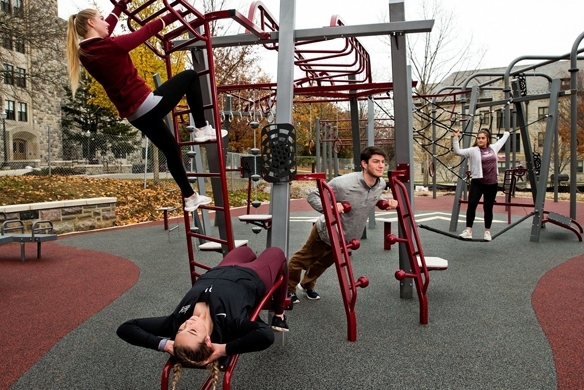 A group of three girls and one guy performing various workouts on outdoor exercise equipment that is on a playground.  