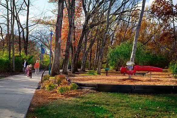 A scenery of a paved walkway going past a playground that has a red dragonfly which people are able to ride on