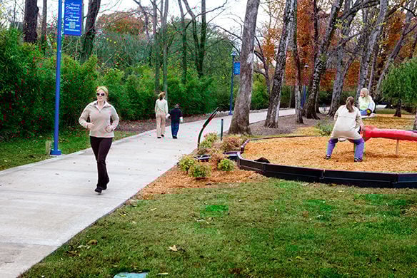 People exercising on a paved walkway as a middle-aged lady and child play on a seesaw beside the people walking