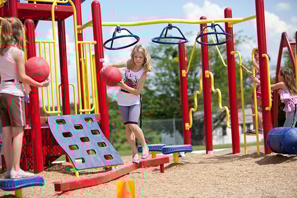 A young girl holding a red ball as she walks across a red balance beam on a playground