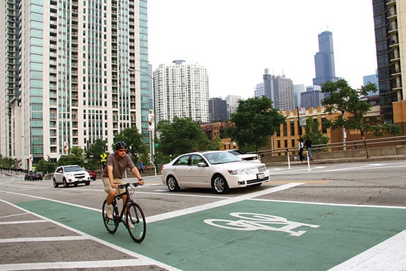 A man riding a bike down a street in a green colored biker's lane. There are also cars beside the person on the road