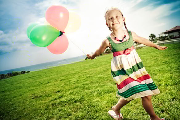 A young girl in a striped dress holding green and pink balloons in a field beside a body of water