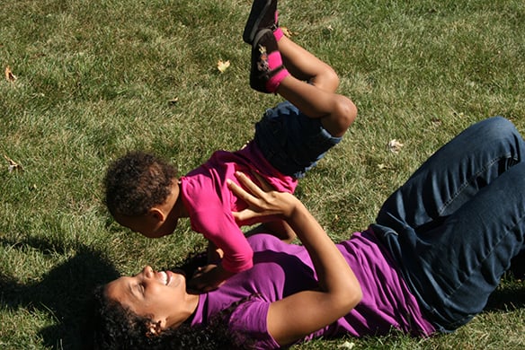 A mother lifting her daughter above her in the air as the mother lies down in the grass