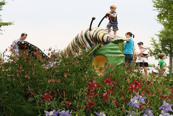 Playground with a large caterpillar sculpture that a child is standing on top of. The playground also contains people, flowers, and greenery nearby.