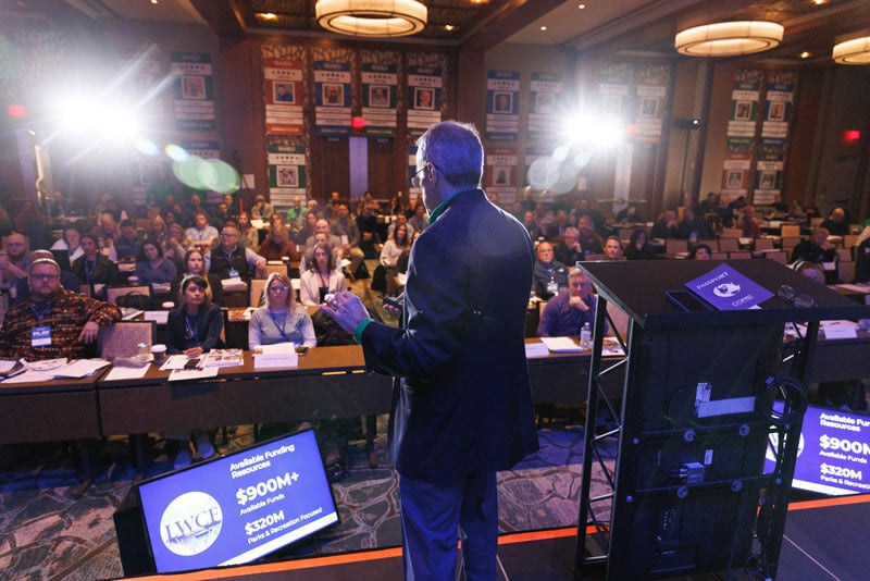 A man in a sports coat talking to a crowd in person. 