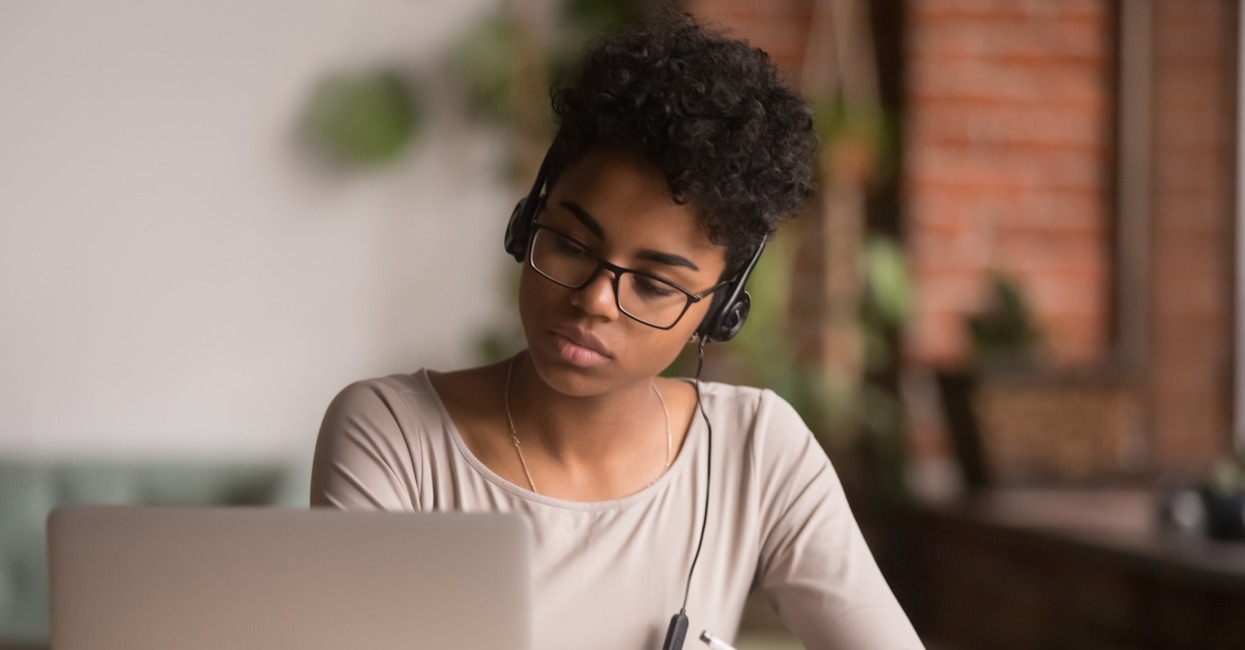 black woman with glasses and headphones sitting in front of a laptop with a pen in hand.