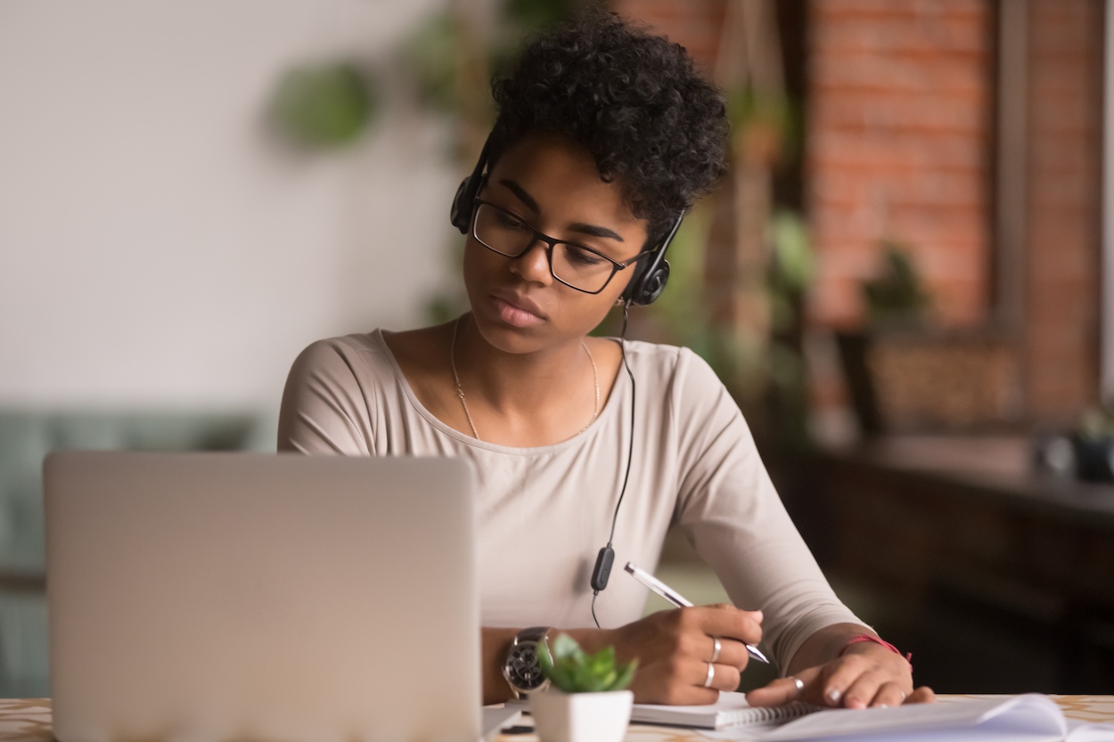 black woman with glasses and headphones sitting in front of a laptop with a pen in hand.