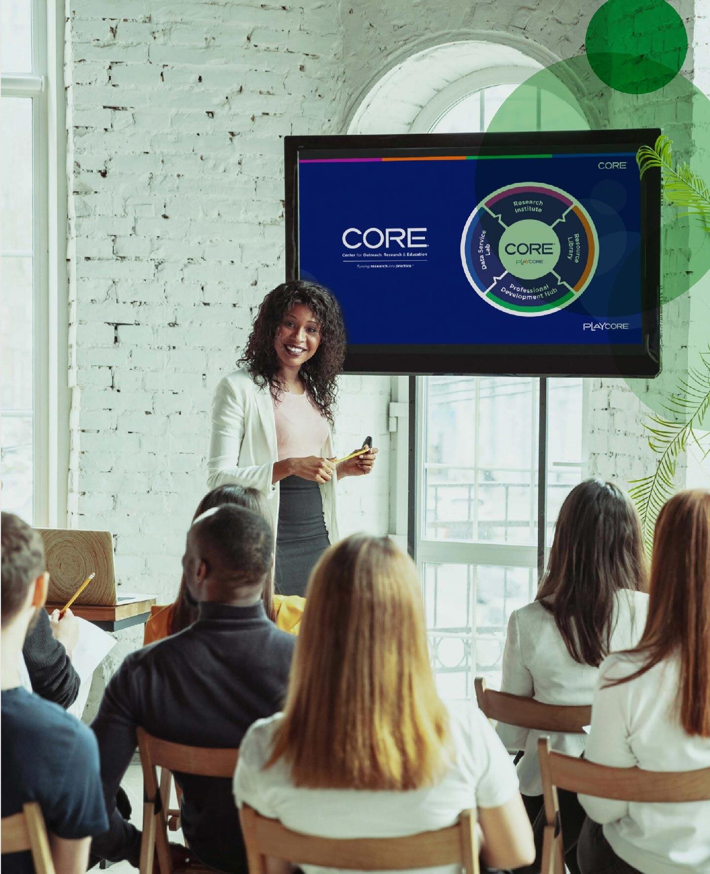 A female speaker talking to a group of people in a room with white bricks. There is also a television with the CORE emblem on it.