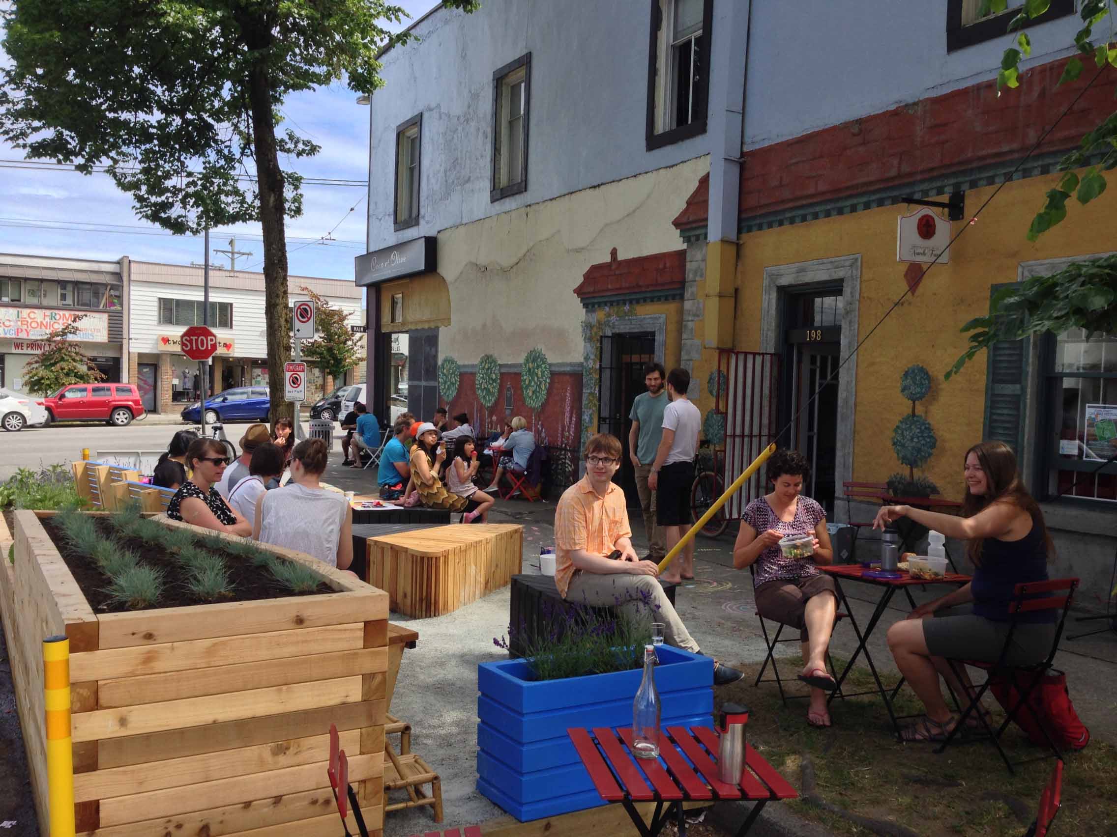 People socializing in colorful urban parklet with seating and planters.
