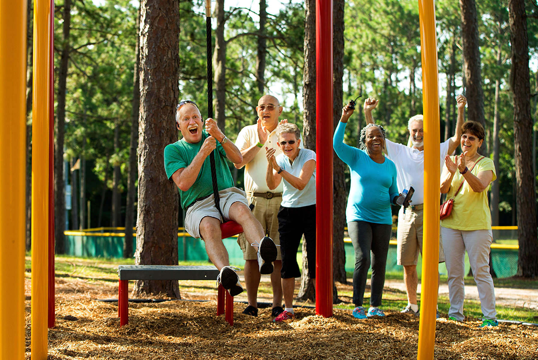 A white-haired man playing on a sliding swing, cheered on by his adult friends