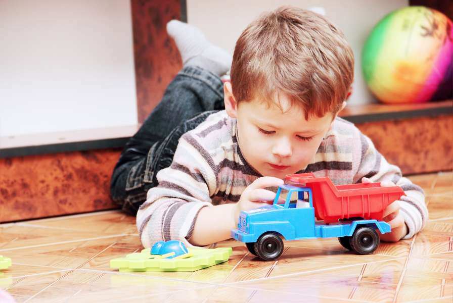 Young boy lying on the floor intently playing with a blue and red toy dump truck, surrounded by other small toys in a cozy indoor setting.