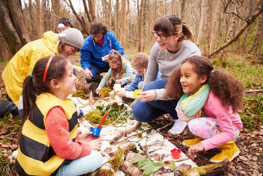 A mixed group of children and adults gathered in a forest around a white tarp covered with various natural products such as twigs, leaves, and moss