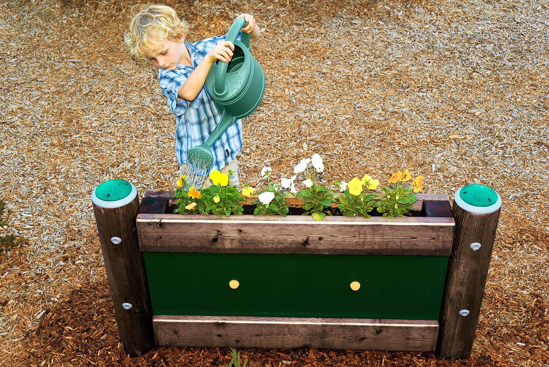 Young child using a green watering can to water colorful flowers in a raised garden bed at a playground, encouraging hands-on learning and sensory play.