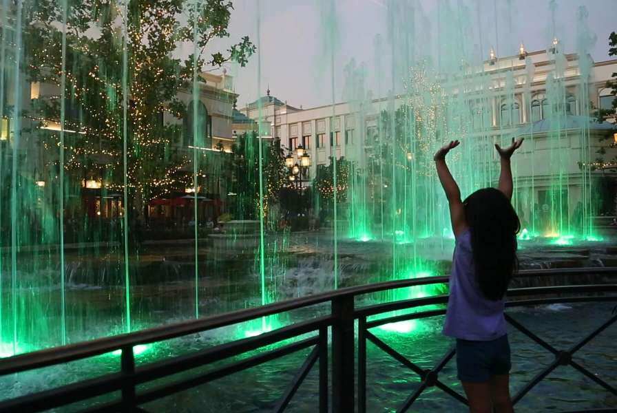 A young girl raising her hands before a water feature illuminated by green lights