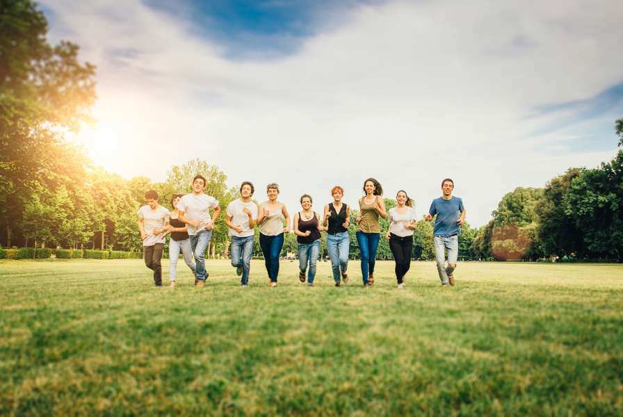 A diverse group of young adults runs joyfully across a wide, grassy field in a sunny park, surrounded by trees and open sky.