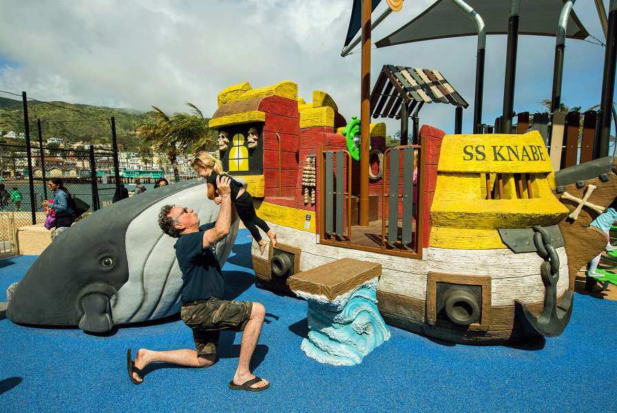 A man raising a child into the air in front front of a playground model of a ship