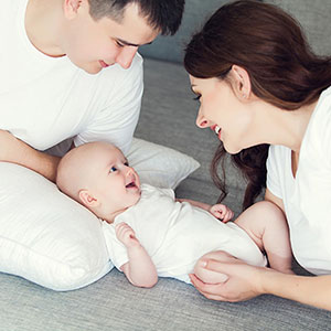 A baby propped up by a pillow being observed by a man and woman, all of whom are wearing white
