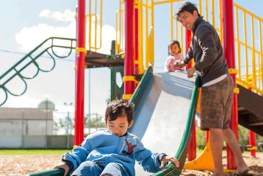 A child at the bottom of a slide being watched over by an adult and followed by a younger child