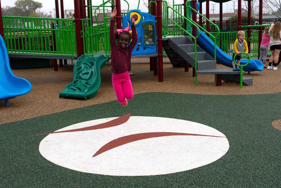 A child wearing a pink bow jumping with a big smile on her face on a playground