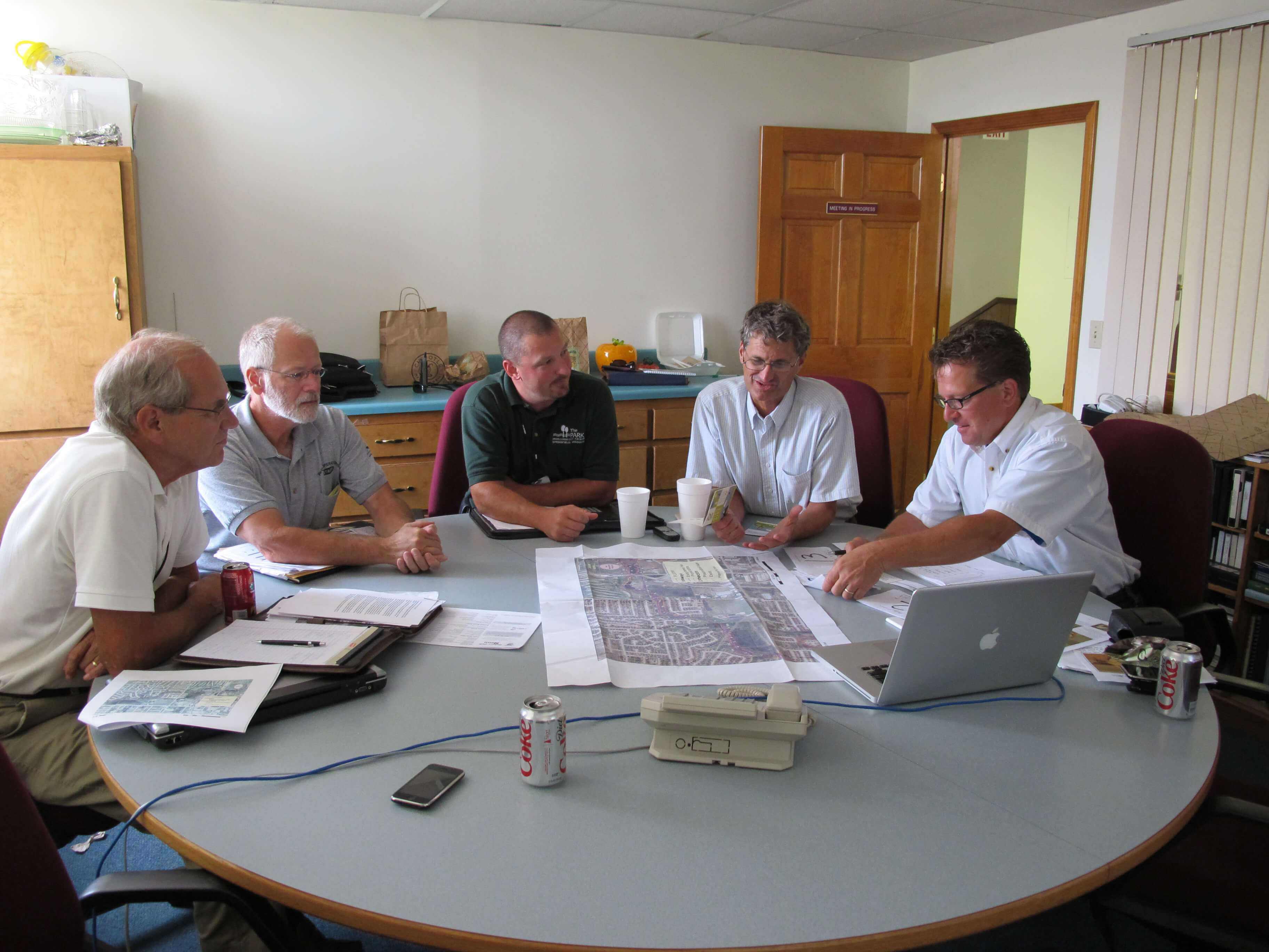 Group of five men in a meeting room reviewing playground or site plans spread across a table, engaged in discussion with laptops, documents, and drinks nearby.