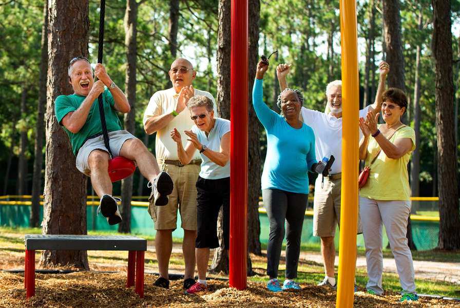 A group of older adults joyfully play at an outdoor park, with one man swinging on a rope swing while others cheer, laugh, and clap in encouragement.