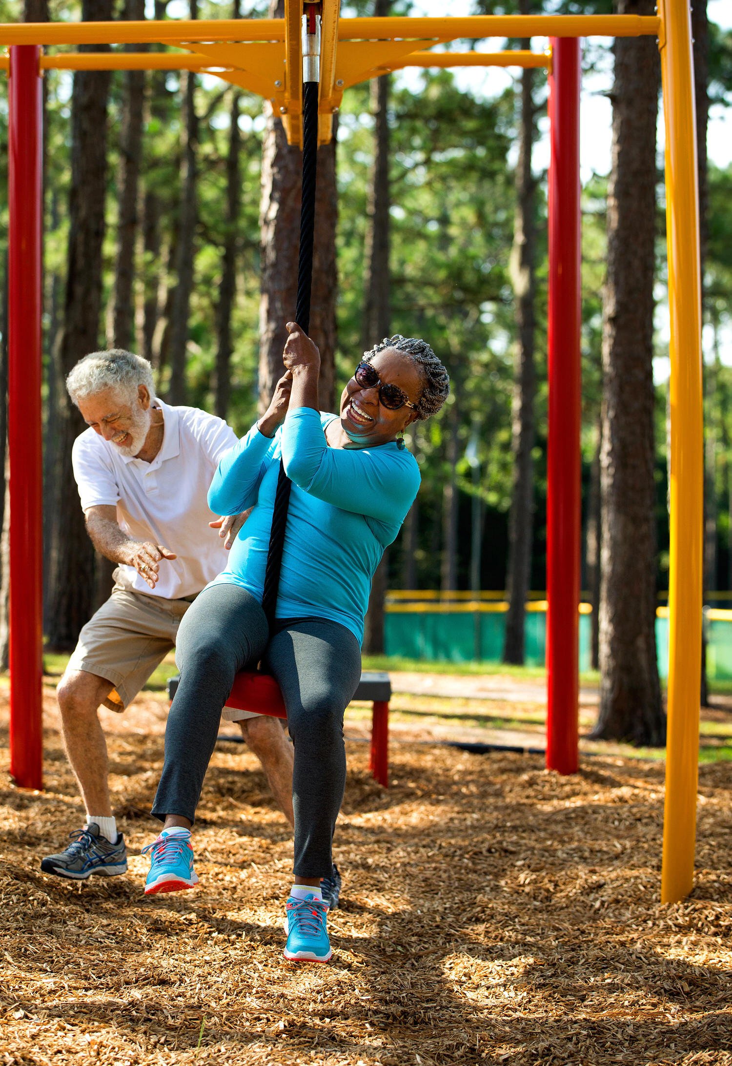 An older woman smiles while riding a zip line swing at an outdoor park, as an older man playfully gives her a push, both enjoying active play in nature.