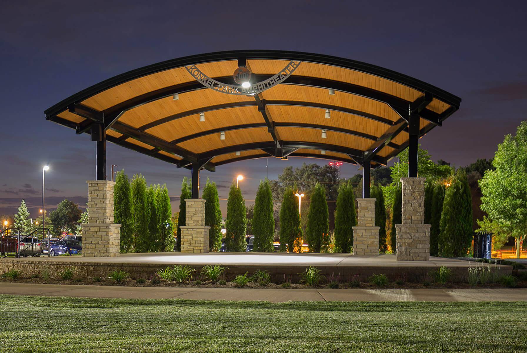 A well-lit pavilion with a curved roof in the evening surrounded by shrubbery