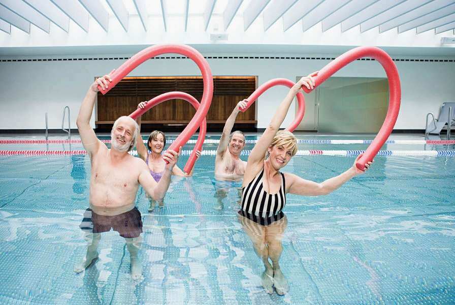 Adults in a swimming pool performing low-impact aerobic exercises with pool noodles raised above their head
